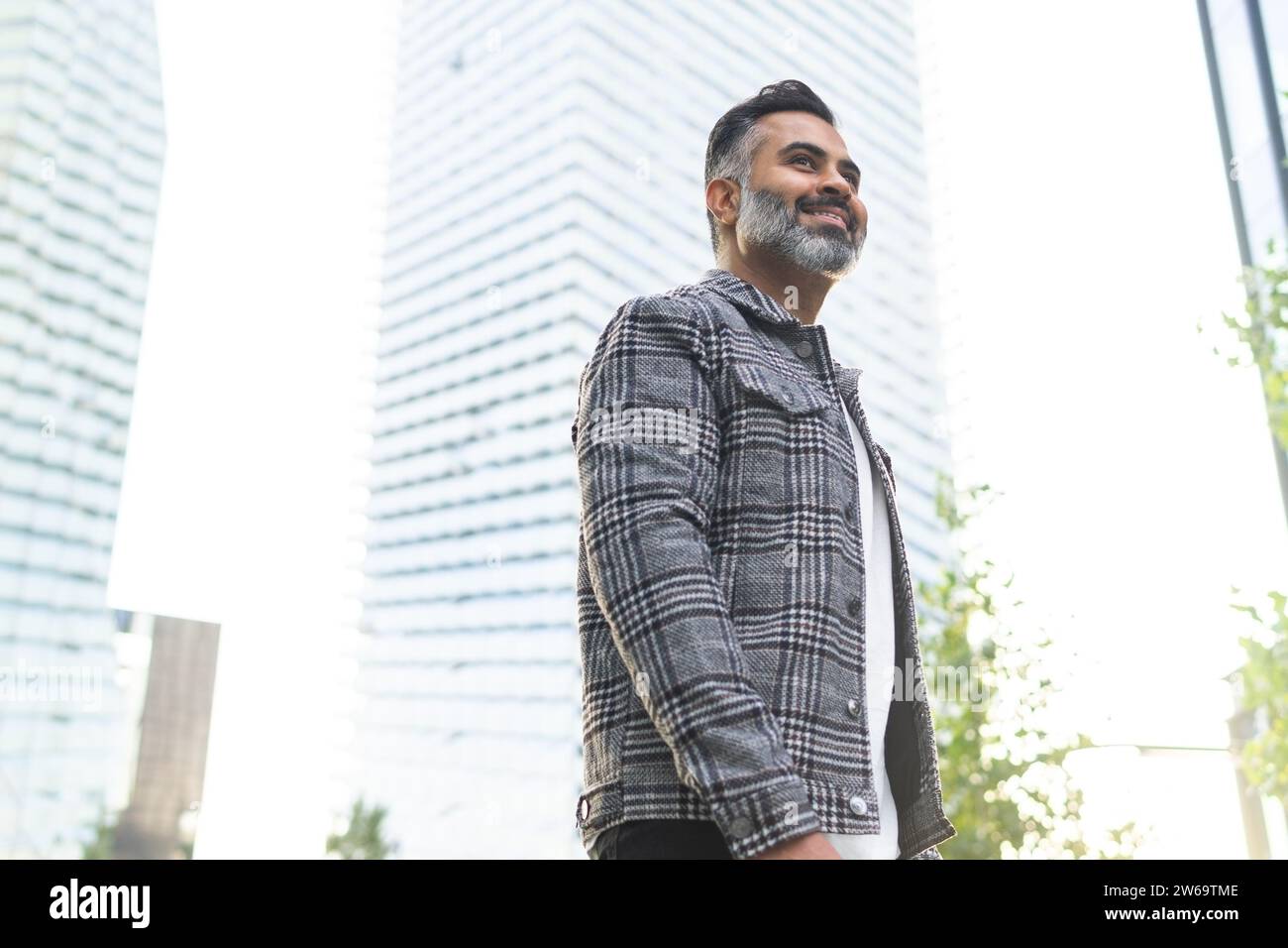 Angolo basso di uomo indiano felice che indossa abiti casual intelligenti passeggiando lungo la strada e sorridendo su un edificio moderno sullo sfondo durante il giorno Foto Stock
