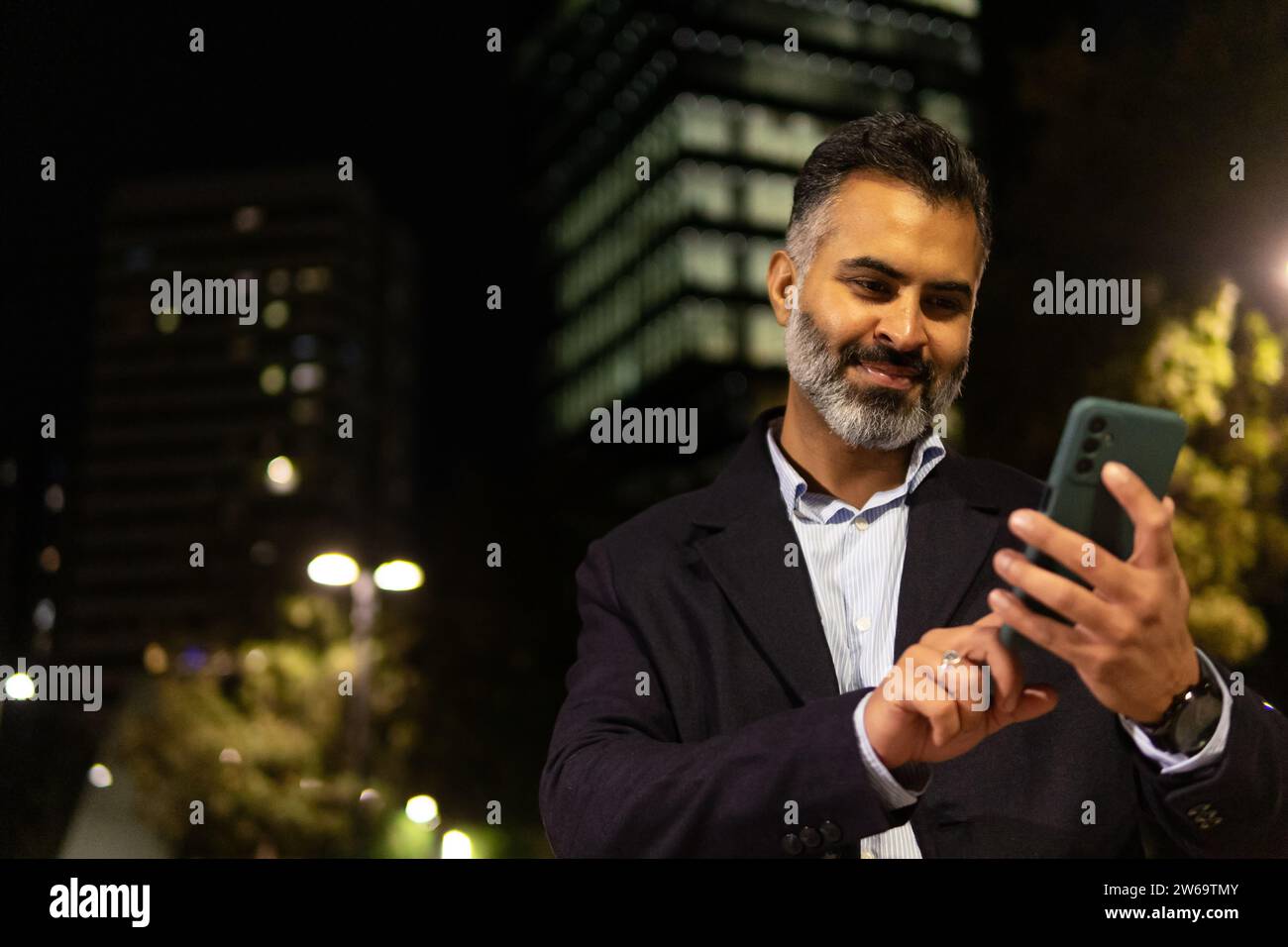 Sorridente uomo indiano barbuto in tuta, in piedi di sera contro lampade ed edifici accesi all'esterno mentre mandavano messaggi con il cellulare Foto Stock