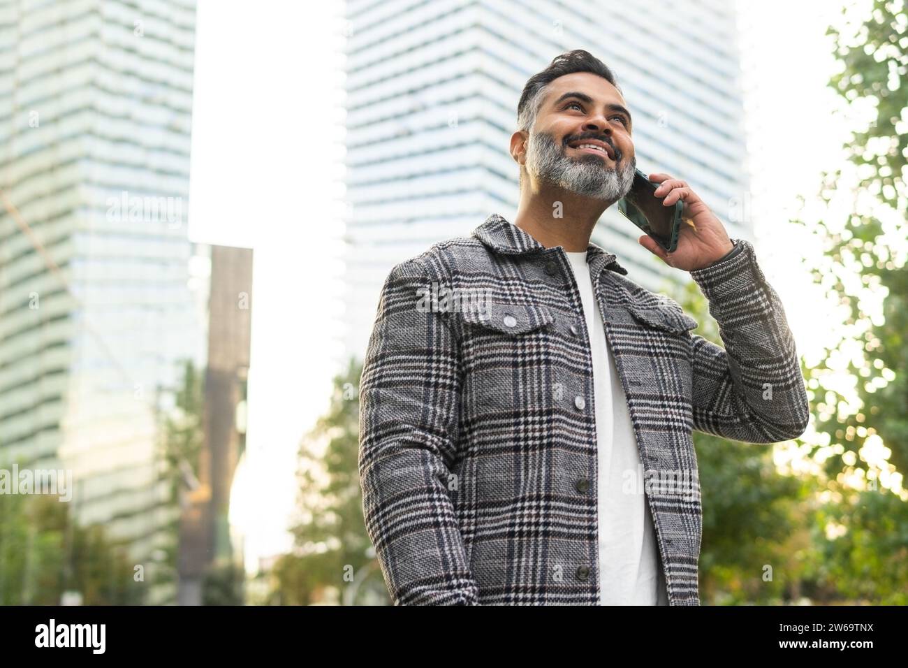 Angolo ridotto di uomo d'affari indiano barbuto positivo con una giacca a scacchi sorridente e parlante su smartphone sullo sfondo sfocato del paesaggio urbano Foto Stock