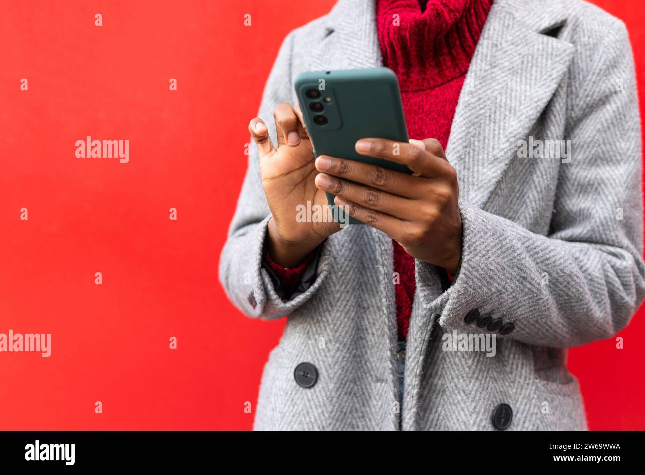Donna afro-americana tagliata e irriconoscibile in uno smartphone da sfogliare mentre si trova in piedi su sfondo rosso Foto Stock