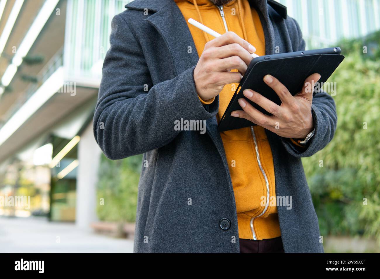 Un professionista aziendale che indossa un abbigliamento casual intelligente utilizza intensamente un tablet digitale che si trova all'esterno di un edificio moderno. Foto Stock