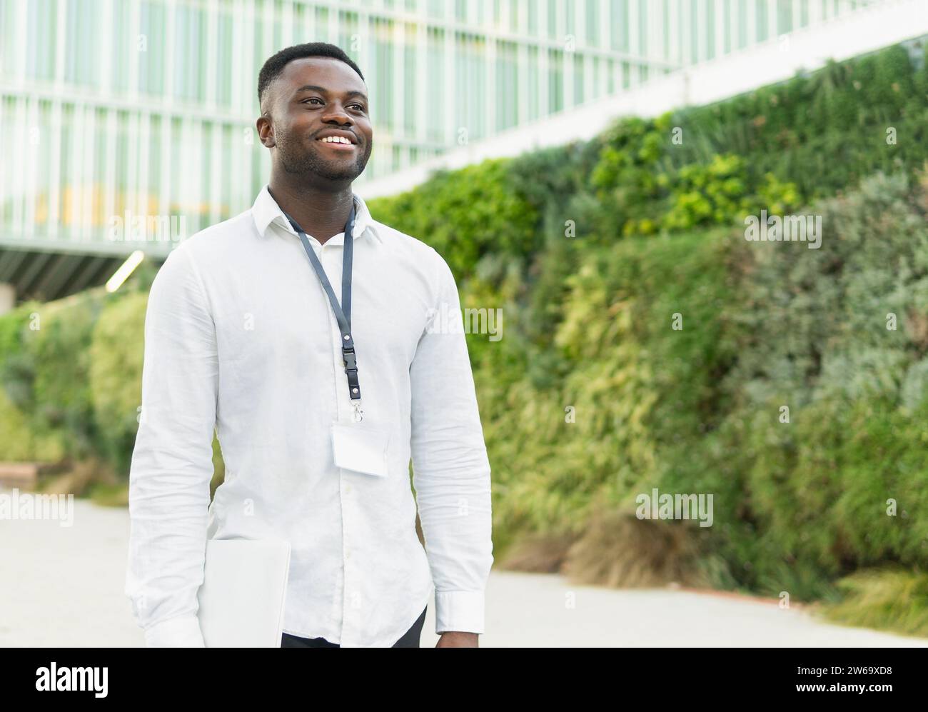 Felice giovane imprenditore afroamericano con camicia bianca che porta netbook mentre cammina fuori dall'edificio di uffici vicino a Hedge sulla strada della città Foto Stock