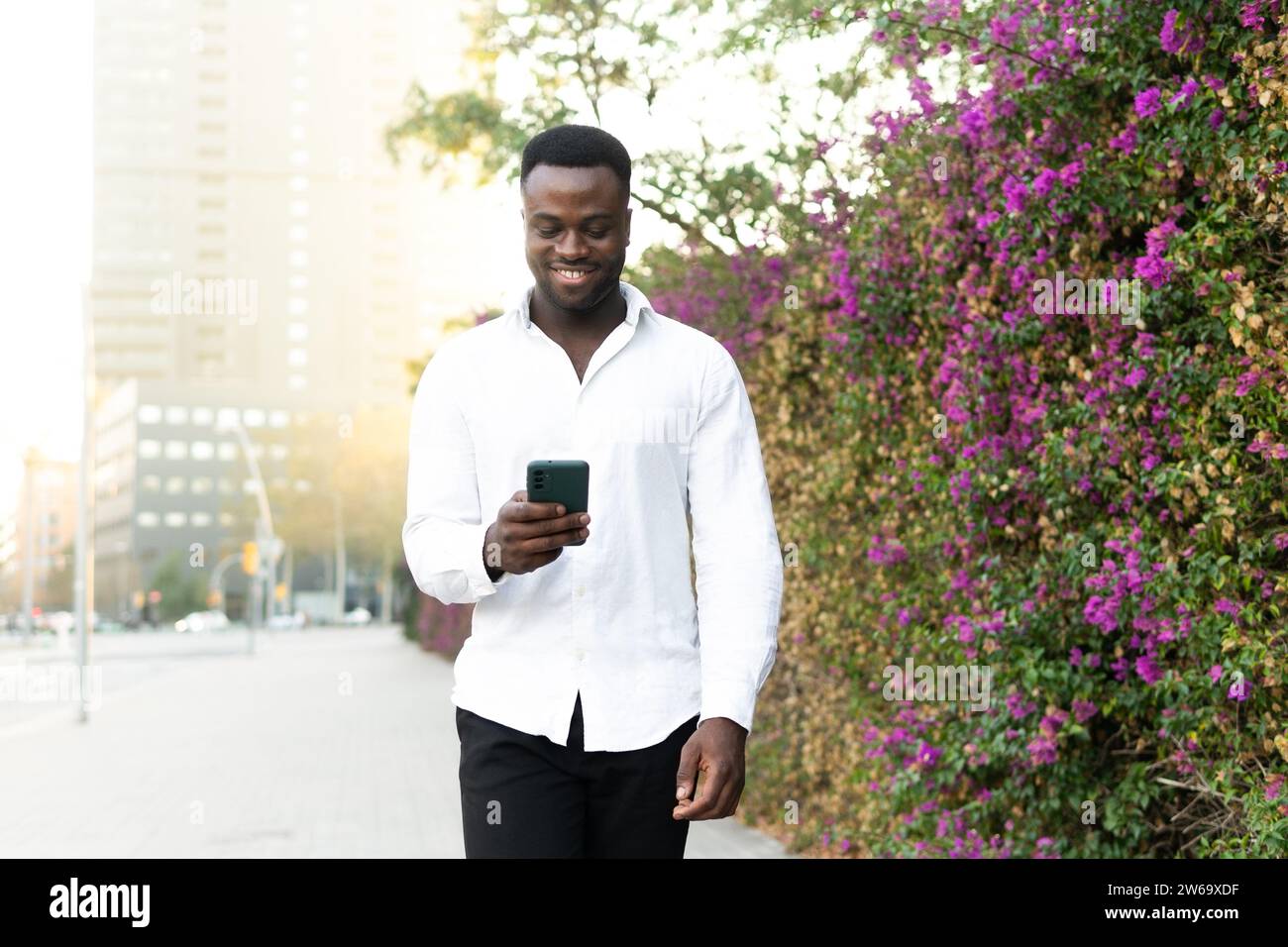 Allegro giovane imprenditore afro-americano con camicia bianca che cammina vicino a cespugli in fiore e usa il telefono cellulare contro gli edifici cittadini sotto il sole Foto Stock
