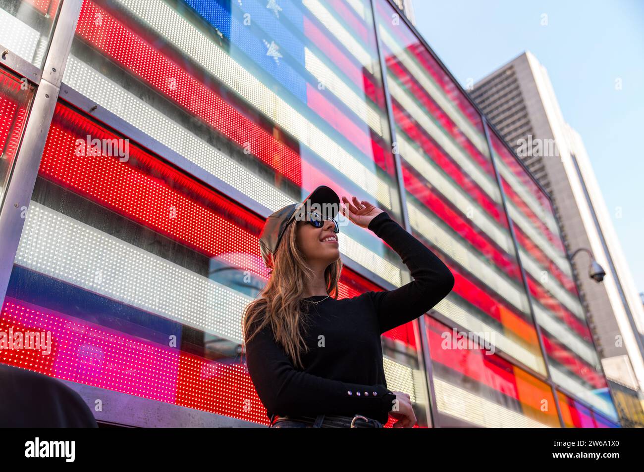 Una giovane donna elegante con occhiali da sole sorride mentre guarda in alto, in piedi di fronte a una vibrante bandiera americana esposta su uno schermo digitale a Manhattan, N. Foto Stock