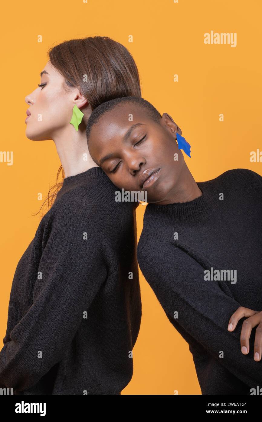 Vista laterale di diverse giovani amiche che si appoggiano l'una all'altra con gli occhi chiusi indossando orecchini colorati alla moda su sfondo giallo Foto Stock