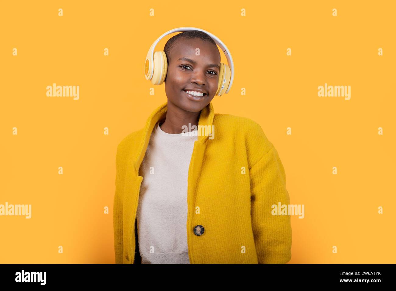 Allegra giovane donna afroamericana che guarda la fotocamera in giacca ascoltando musica attraverso cuffie wireless su sfondo giallo Foto Stock