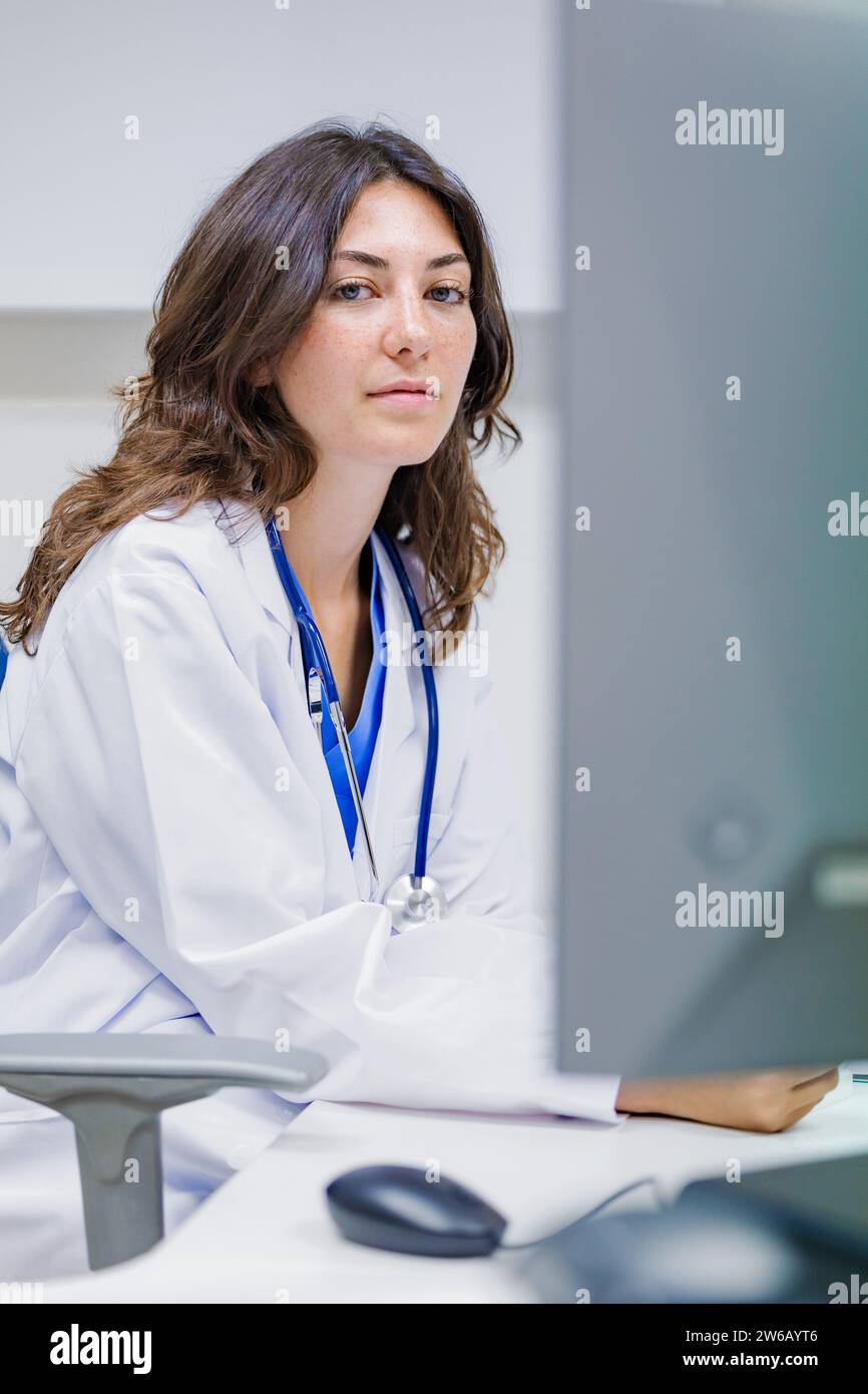 Giovane medico donna che indossa uniforme medica e stetoscopio seduto in ospedale su sfondo bianco mentre guarda la macchina fotografica Foto Stock