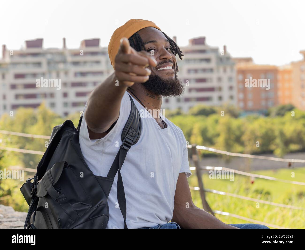 Sorridente giovane hipster afro-americano con cappello giallo con zaino seduto contro gli edifici della città e puntato verso di te Foto Stock