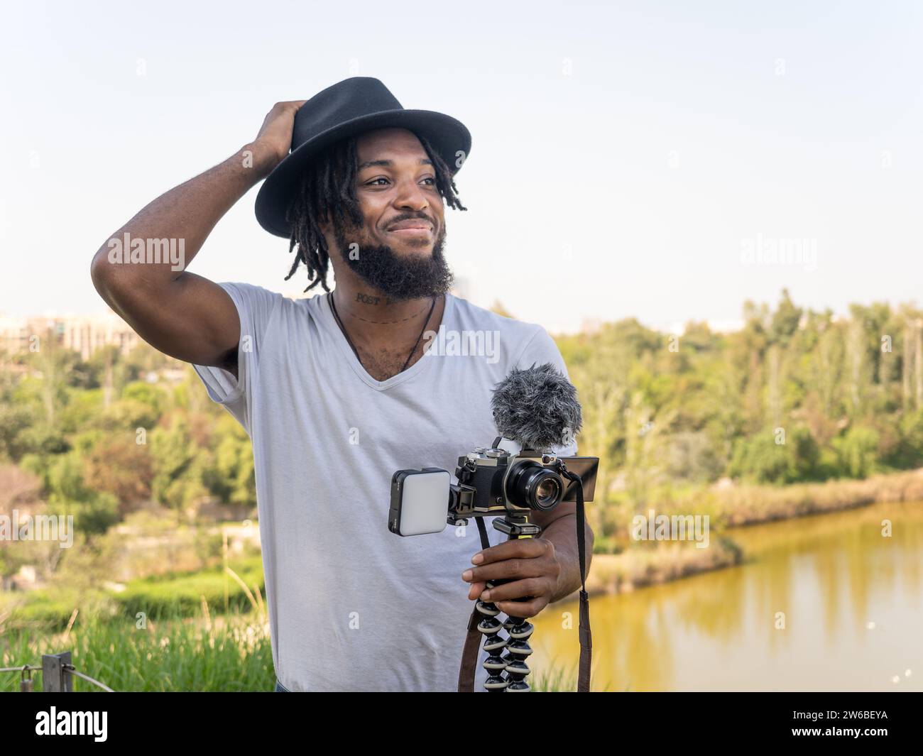 Giovane blogger afro-americano che indossa un cappello in piedi contro il cielo senza nubi e tiene una videocamera professionale con microfono mentre si prepara per le riprese Foto Stock