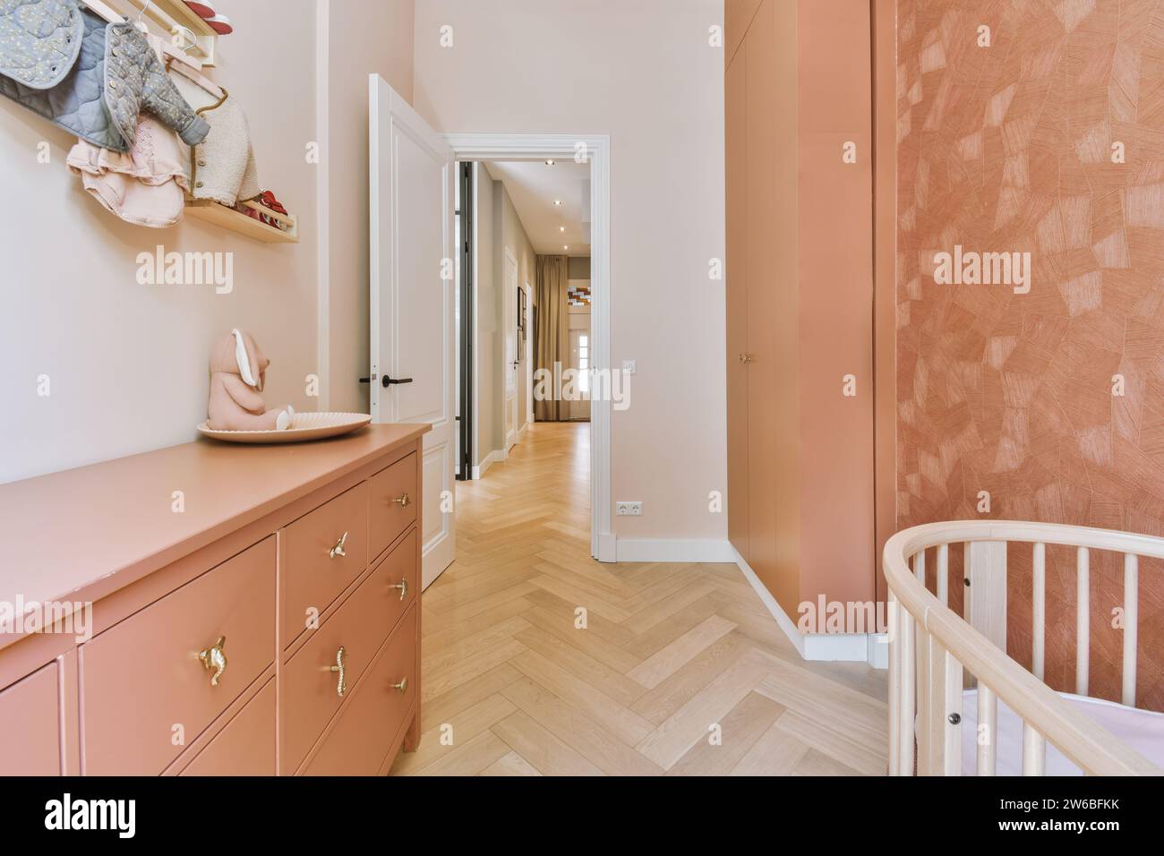 Culla con comò e guardaroba in una stanza rosa per bambini con porta aperta che conduce al corridoio in un appartamento contemporaneo Foto Stock