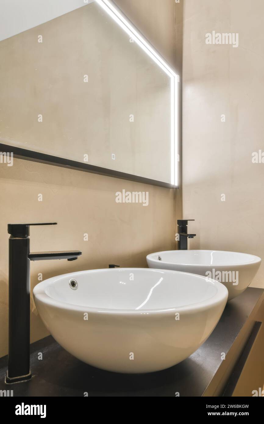 Un bagno elegante e moderno con due lavandini bianchi con eleganti rubinetti neri, sotto una luce a specchio geometrica a LED. Foto Stock