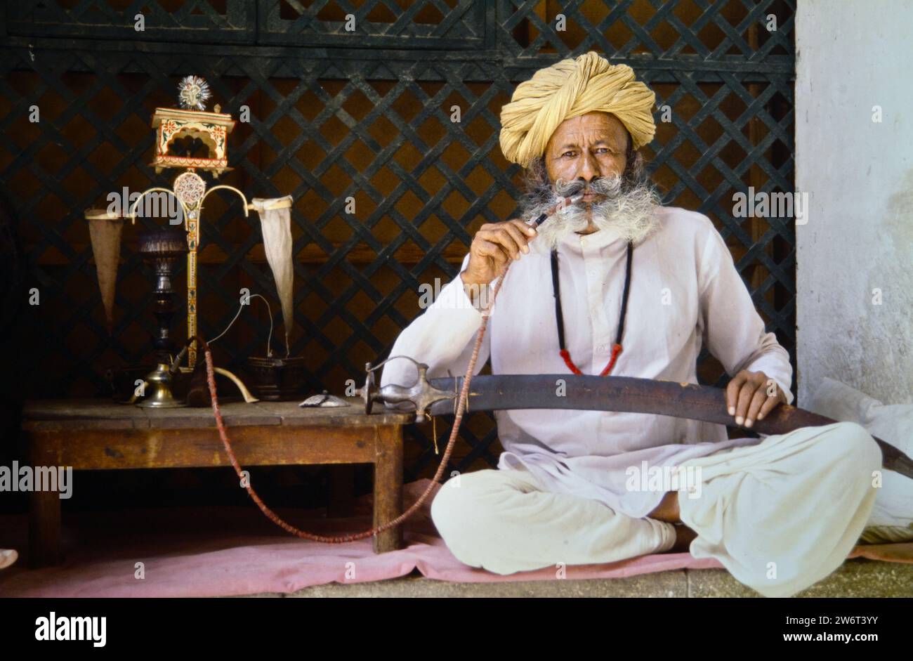 Archivio immagine storica di Un uomo rajasthano con barba, baffi in colorato Turban sitting Cross Legged Smoking A Hookah Pipe Holding A Talwar Curved Sword with Knuckle Bow in A Leather Sheath, Jaisalmer, Rajasthan, India 1990 Foto Stock