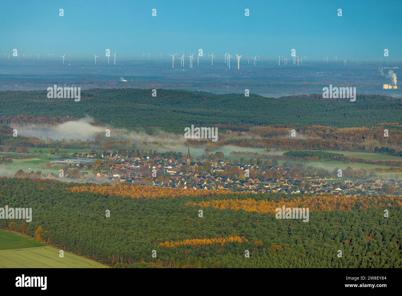 Vista aerea, nebbia sul villaggio di Hullern con St Andreas Church, parco delle turbine eoliche sullo sfondo, circondato da alberi decidui autunnali, Hu Foto Stock