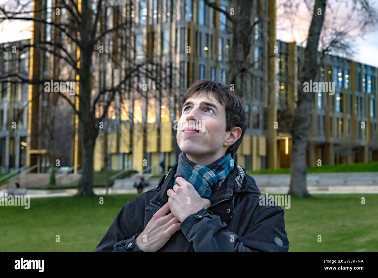 Un giovane in piedi di fronte a un edificio universitario in una giornata fredda, che fissa la sciarpa e sorride guardando verso l'alto e forse verso il futuro. Foto Stock