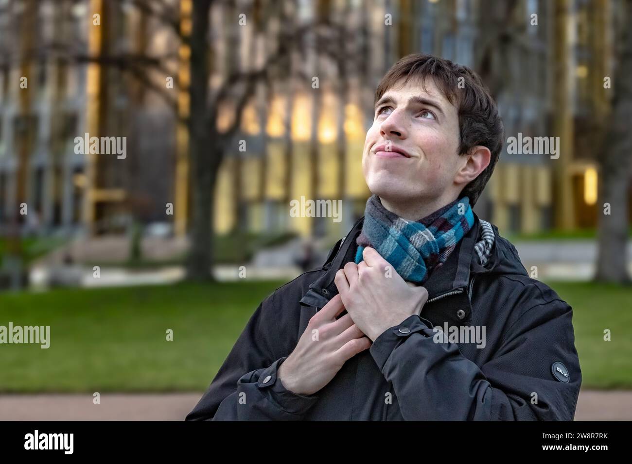 Un giovane in piedi di fronte a un edificio universitario in una giornata fredda, che fissa la sciarpa e sorride guardando verso l'alto e forse verso il futuro. Foto Stock
