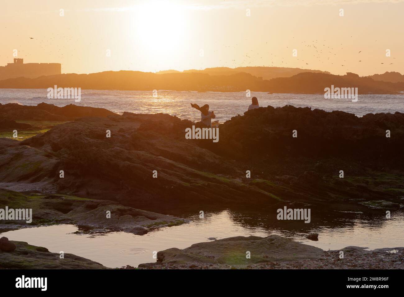 Le persone si siedono sulle formazioni rocciose guardando il tramonto sul mare a Essaouira, la "città ventosa", in Marocco. 21 dicembre 2023 Foto Stock