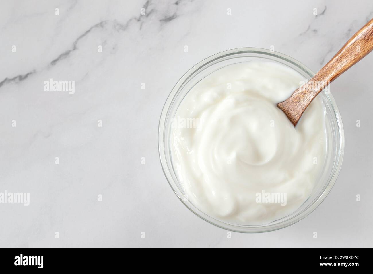 Yogurt greco in una ciotola di vetro con cucchiaio di legno su sfondo bianco. Vista della tabella superiore. Copia spazio. Cibo proteico sano. Foto Stock