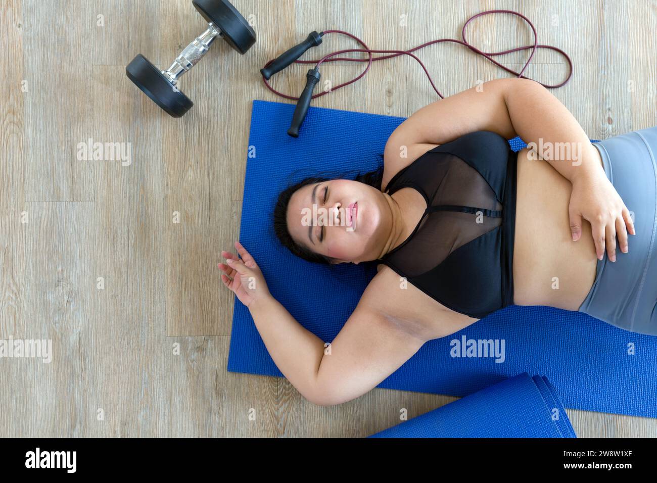 Una donna più grande si trova sfinita su un tappetino blu e si prende una pausa dopo una vigorosa sessione di allenamento con manubri e corda da salto. Vista dall'alto Foto Stock