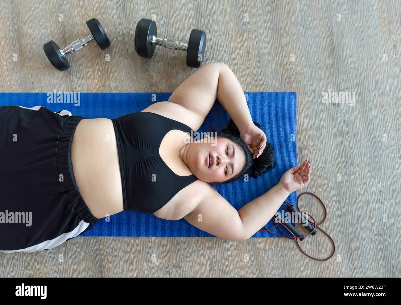 Una donna più grande si trova sfinita su un tappetino blu e si prende una pausa dopo una vigorosa sessione di allenamento con manubri e corda da salto. Vista dall'alto Foto Stock