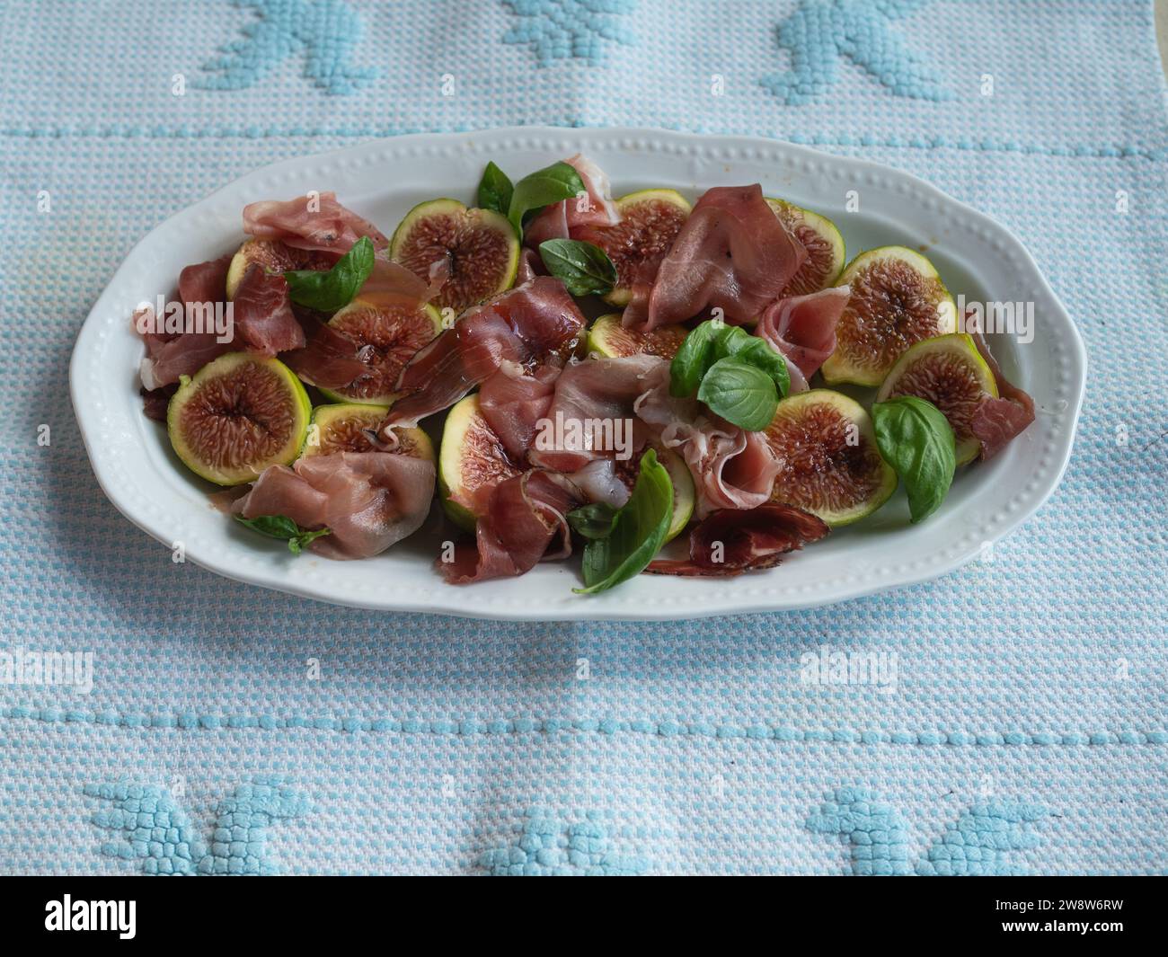 Orizzontalmente direttamente dall'alto, ripresa ravvicinata di insalata di fichi e prosciutto adornata con foglie di basilico fresco servita su un delicato piatto bianco su un tradizionale Foto Stock