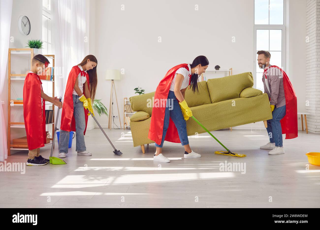Felice famiglia sorridente con bambini in costumi da supereroi che puliscono il pavimento insieme a casa. Foto Stock