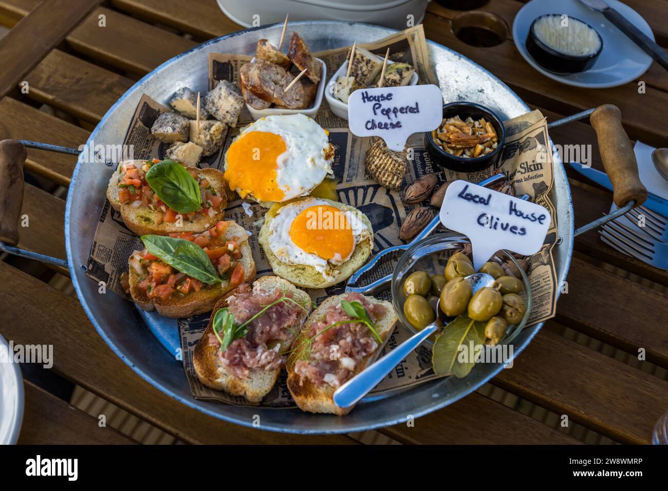 Un vassoio condiviso alla fattoria turistica Tuta a Kerċem, Gozo, Malta, combina formaggio al pepe fatto in casa, olive prodotte in casa, bruschetta al pomodoro e basilico, uova fritte e una tartara di carne su pane tostato. Piccoli cartoncini etichettano il "Formaggio al pepe casa" e le "Olive locali casa". Foto Stock