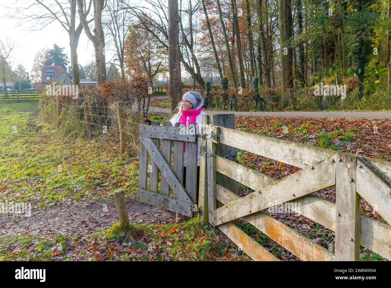Donna adulta anziana che entra nella riserva naturale olandese di Geleenbeekdal, cancello con recinzione in legno, alberi nudi sullo sfondo, giacca bianca, scialle rosa, giorno a Scaligna Foto Stock