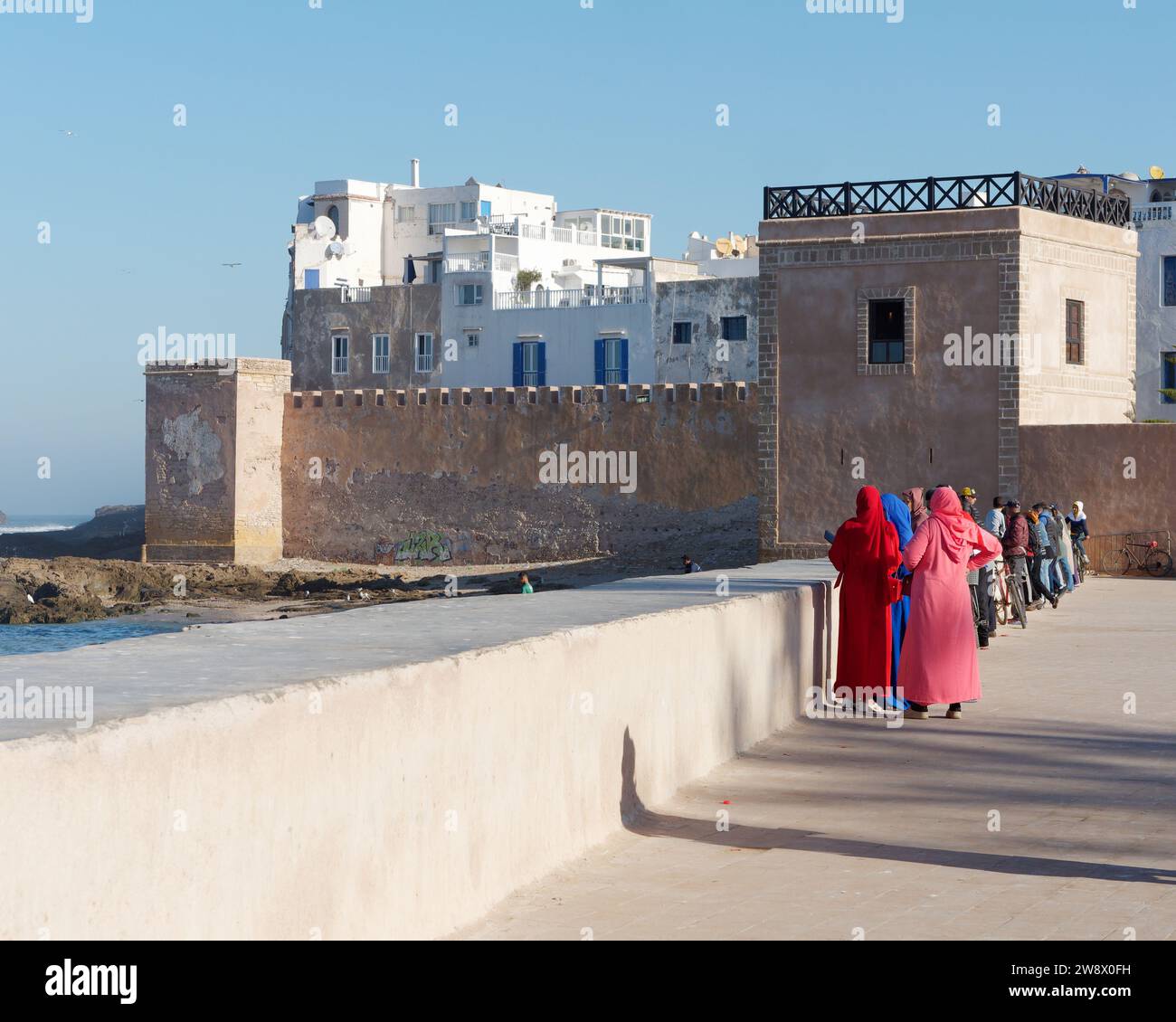 Le donne in abiti colorati si trovano accanto alle mura della città con la costa e gli edifici della medina alle spalle. Essaouira, Marocco. 22 dicembre 2023 Foto Stock