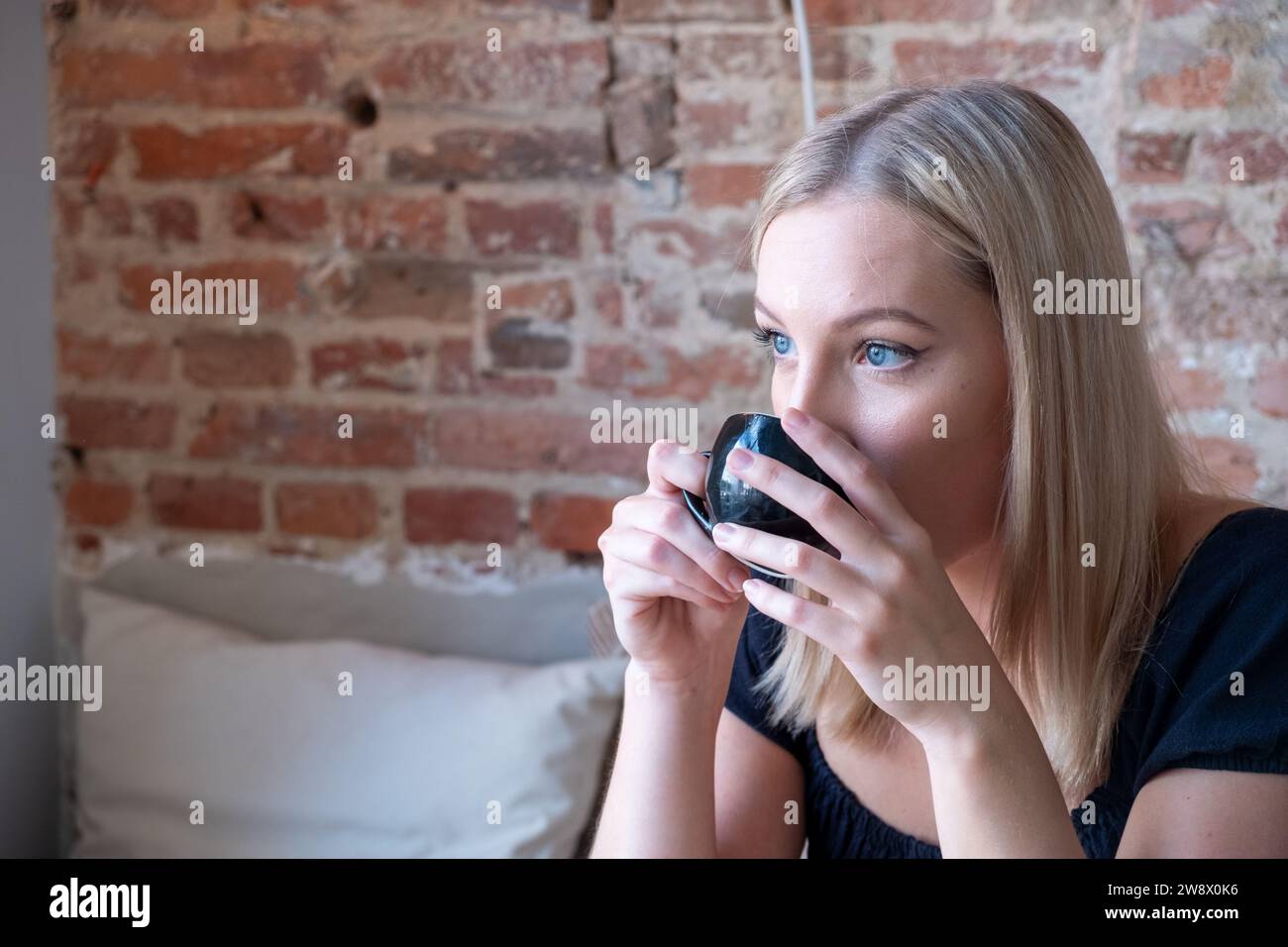 L'immagine ravvicinata cattura una giovane donna con i capelli biondi mentre sorseggia un sorso da una piccola tazza di caffè e i suoi occhi guardano lontano. E' seduta in un angolo intimo di un caffe' che vanta un fascino rustico, con un muro di mattoni a vista che funge da sfondo. La natura intima della fotografia suggerisce un momento di relax e divertimento, mentre si prende una pausa nella sua giornata per assaporare il caffè e l'ambiente circostante. Primo piano di Young Woman che sorseggia un caffè in un accogliente Cafe Corner. Foto di alta qualità Foto Stock