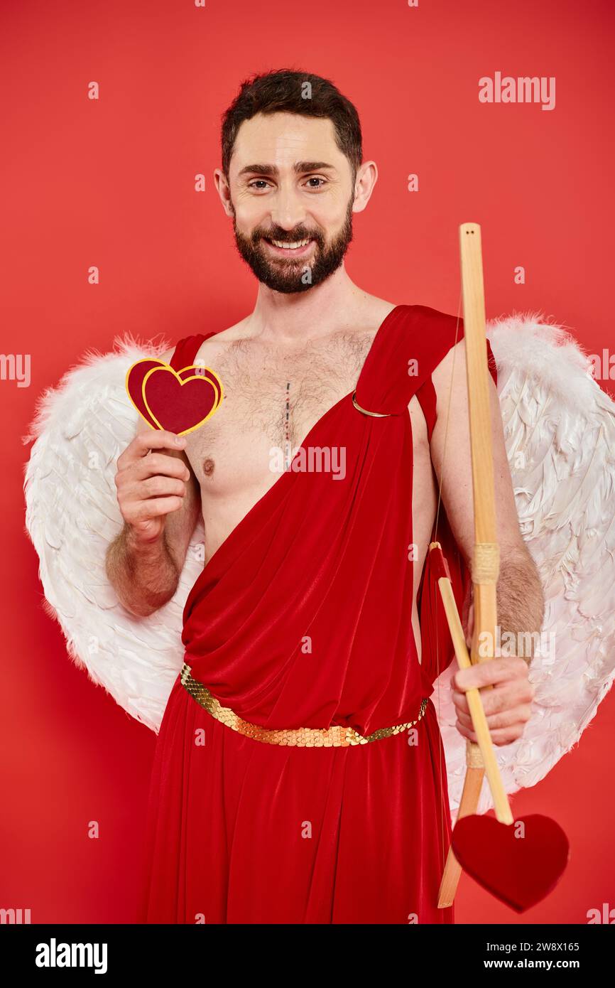 felice uomo barbuto in costume di cupido con cuori di carta e arco con freccia sul rosso, giorno dei san valentini Foto Stock