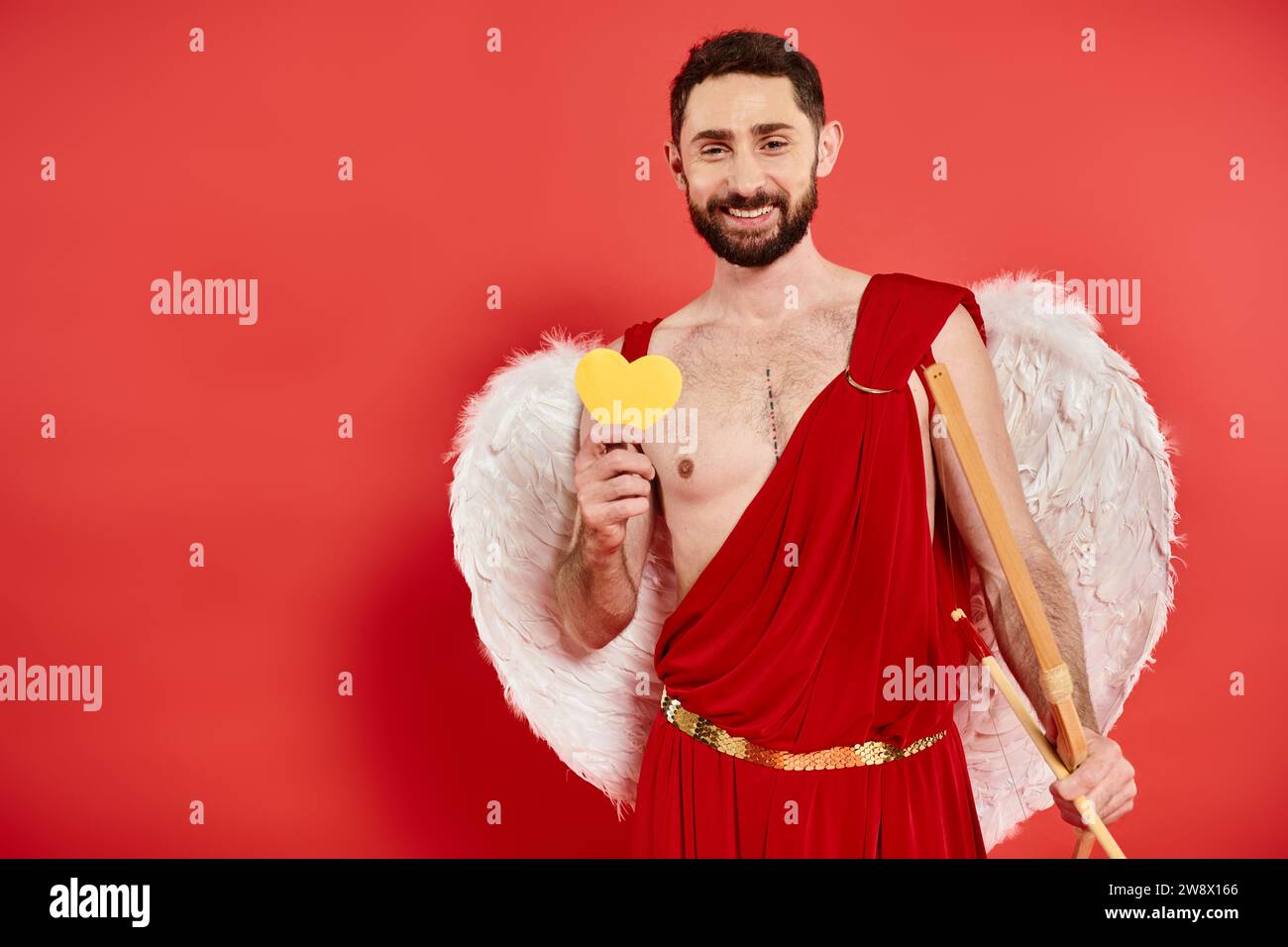 Allegro uomo barbuto in costume cupido con fiocco e cuore di carta gialla in rosso, giorno di San Valentino Foto Stock