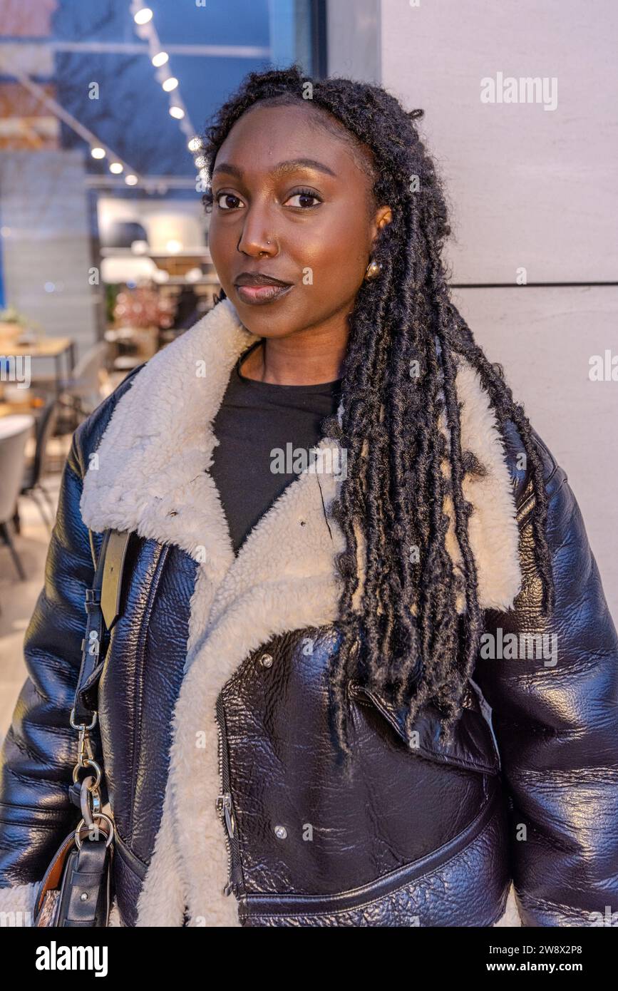 Un'elegante donna africana si trova in un ambiente urbano durante la sera. È vestita alla moda con una giacca in pelle nera con colletto in shearling bianco, che si abbina ai suoi lunghi dreadlocks contorti. Lo sfondo presenta una caffetteria con luce calda che illumina delicatamente il suo viso e le luci a corda aggiungono un effetto bokeh alla scena. Il trucco della donna è fatto con gusto, mettendo in evidenza la sua bellezza naturale e il suo comportamento pronto. Il suo sguardo è diretto verso la telecamera con un sorriso leggero e sicuro, suggerendo sia la facilità di accesso che l'autosicurezza. Urban Chic Black Girl. Alta qualità Foto Stock