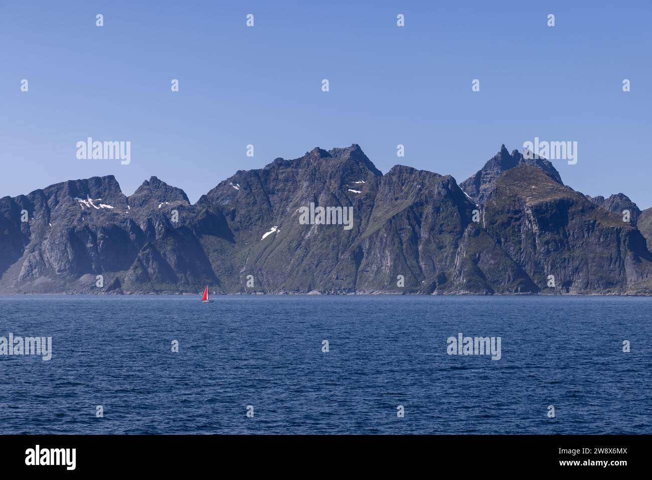 Una vista pittoresca del Mare del Nord e una cresta di montagna che torreggia su acque calme, con una barca a vela che porta una vela rossa vicino all'Isola di Lofoten, sotto un bl Foto Stock