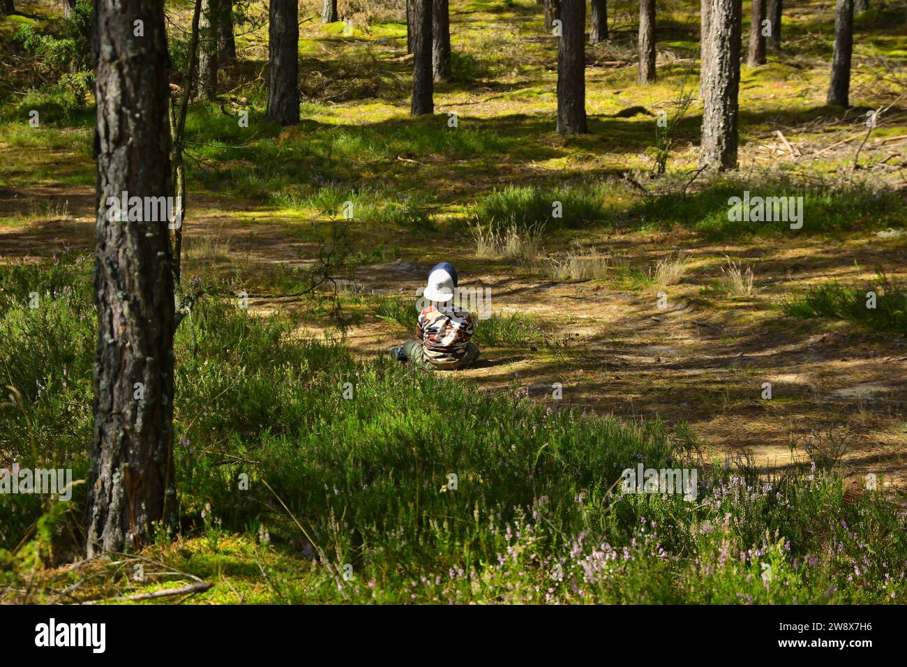 Un bambino in uniforme mimetica siede tutto solo nel mezzo della foresta. dintorni bellissimi, erba e alberi Foto Stock