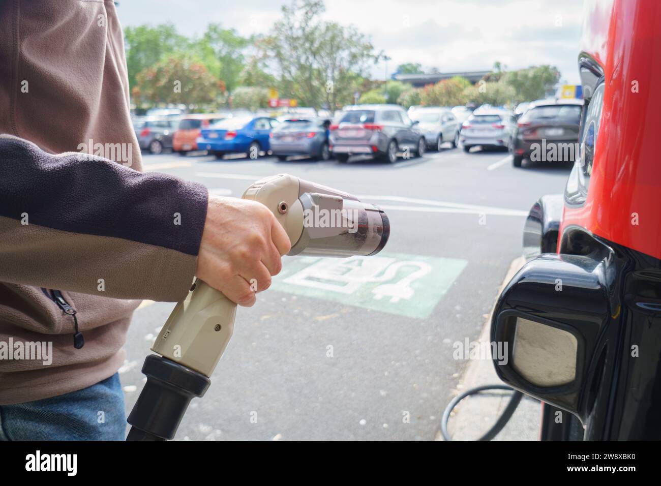 Tenere a mano un caricabatterie per auto elettriche presso un parcheggio con il cartello di ricarica EV a terra. Auto non riconoscibili parcheggiate sullo sfondo. Foto Stock