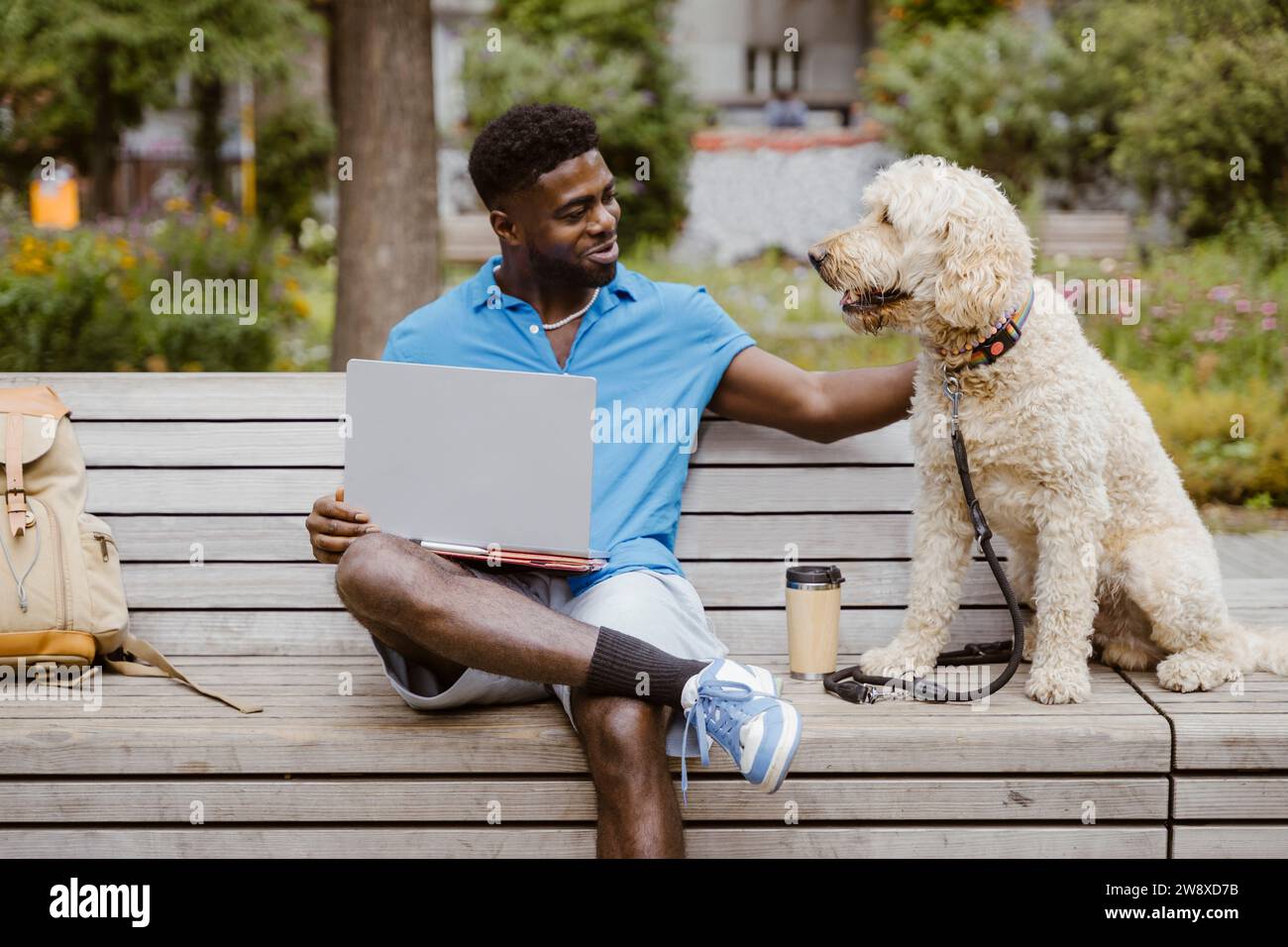 Un giovane sorridente con un computer portatile che accarezza il cane seduto sulla panchina nel parco Foto Stock