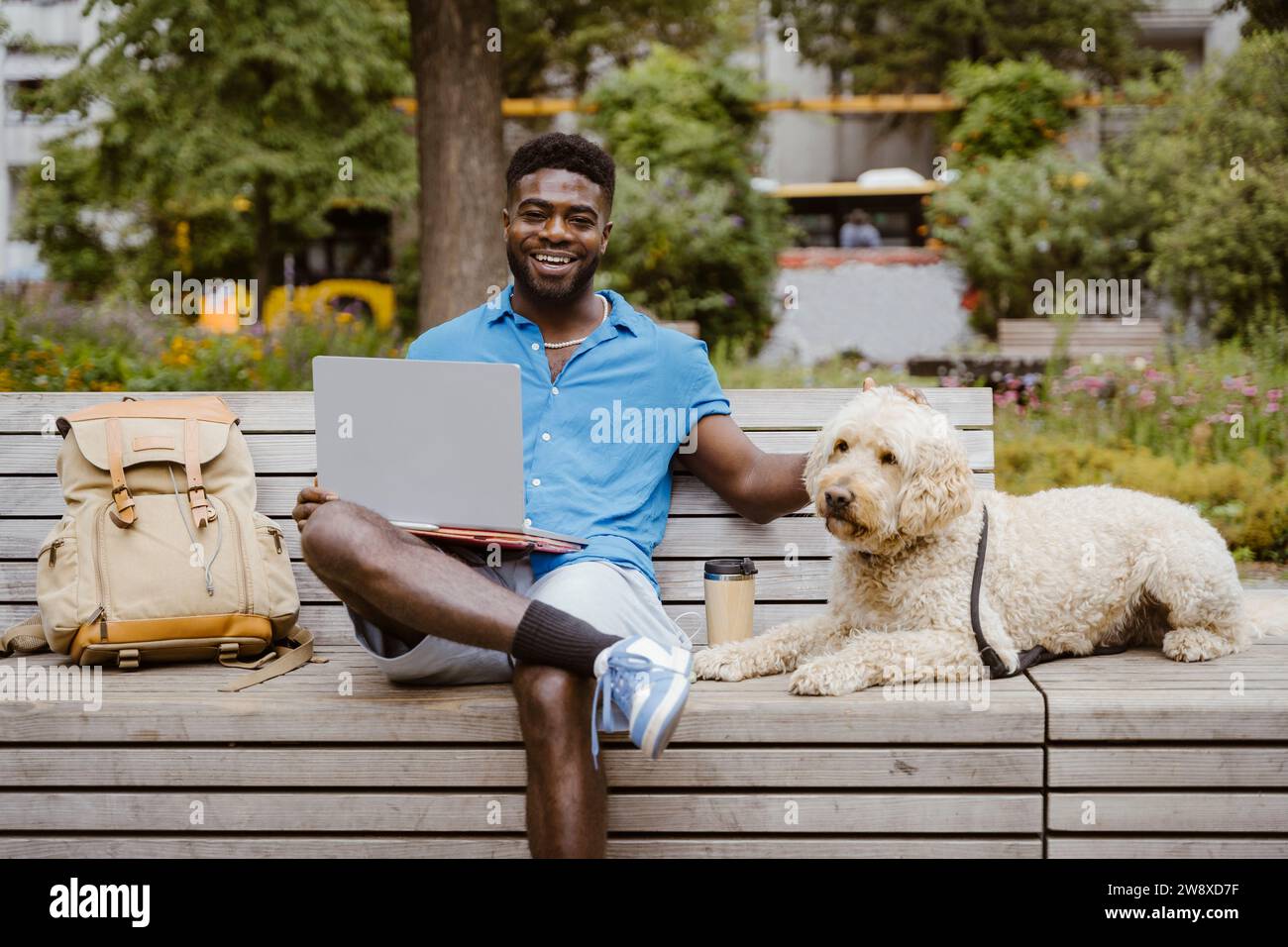 Ritratto di un uomo sorridente con un computer portatile seduto accanto a un cane labradoodle sulla panchina nel parco Foto Stock