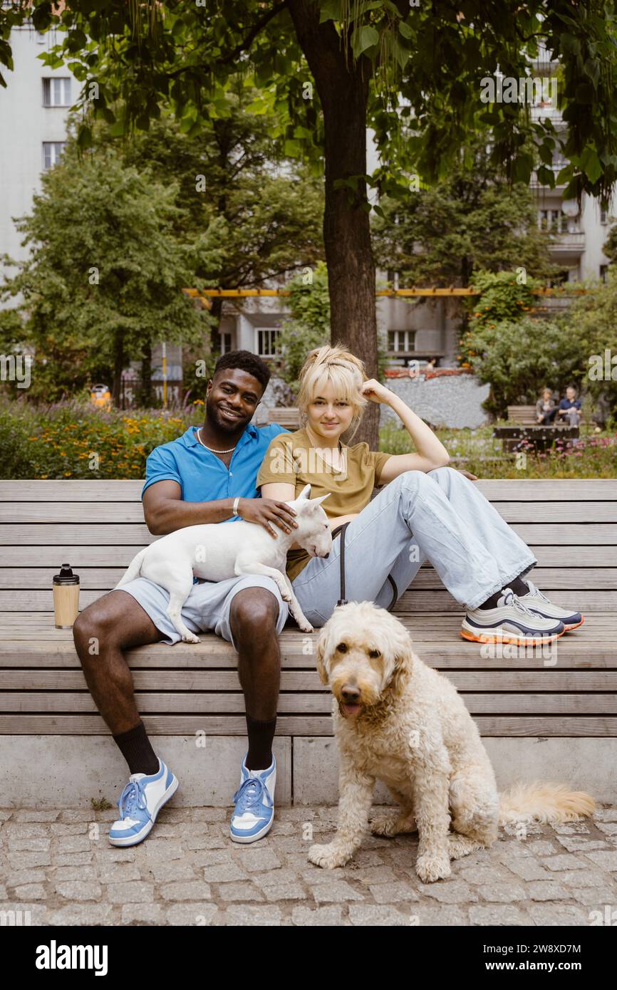 Ritratto di un uomo e una donna sorridenti seduti con cani sulla panchina nel parco pubblico Foto Stock
