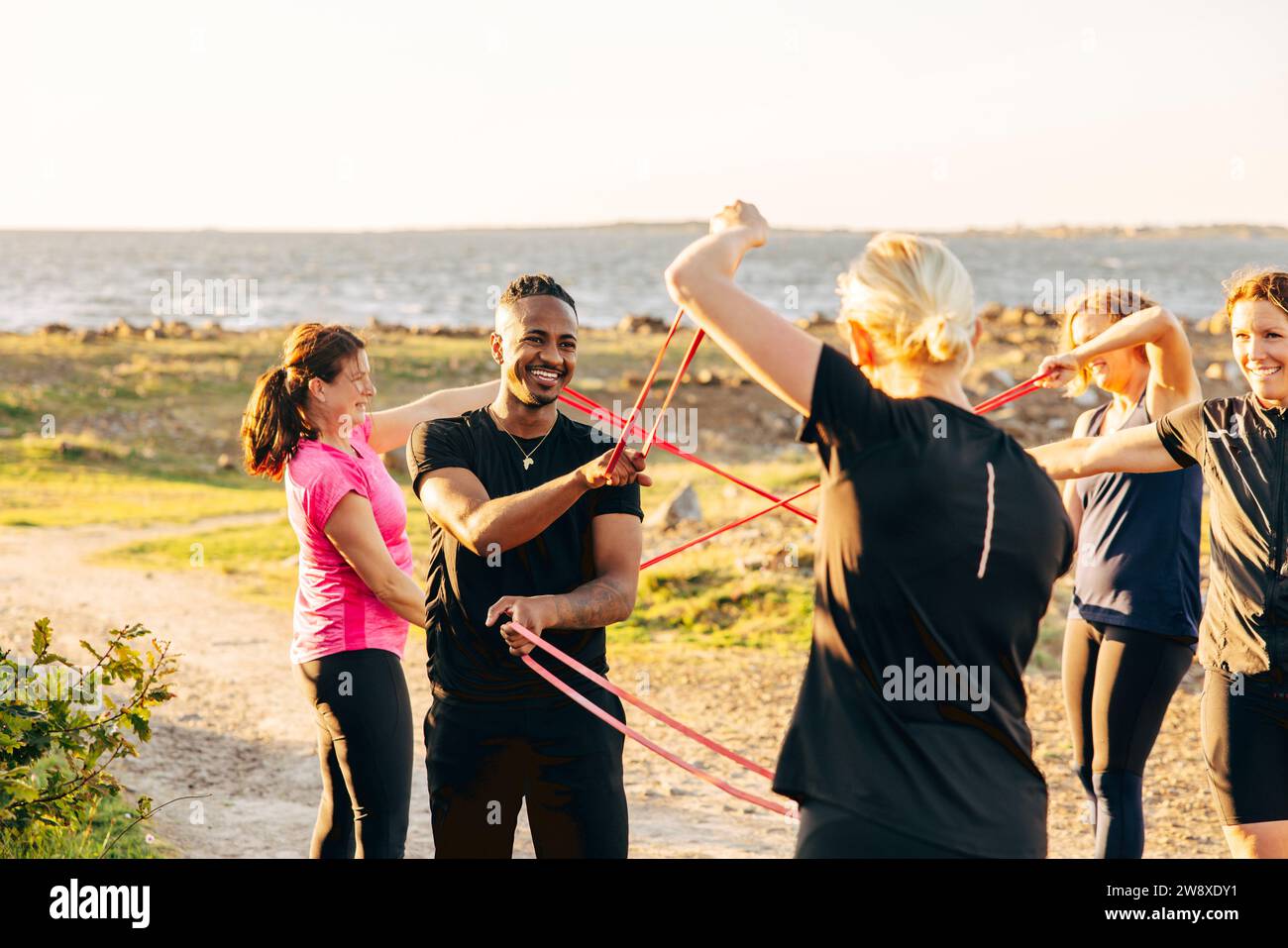 Sorridenti compagni di squadra maschili e femminili che si allenano insieme alle bande di resistenza durante la sessione di allenamento di gruppo in spiaggia Foto Stock