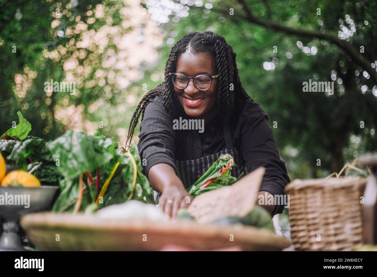 Fornitore femminile sorridente con capelli intrecciati che organizza verdure sul mercato Foto Stock