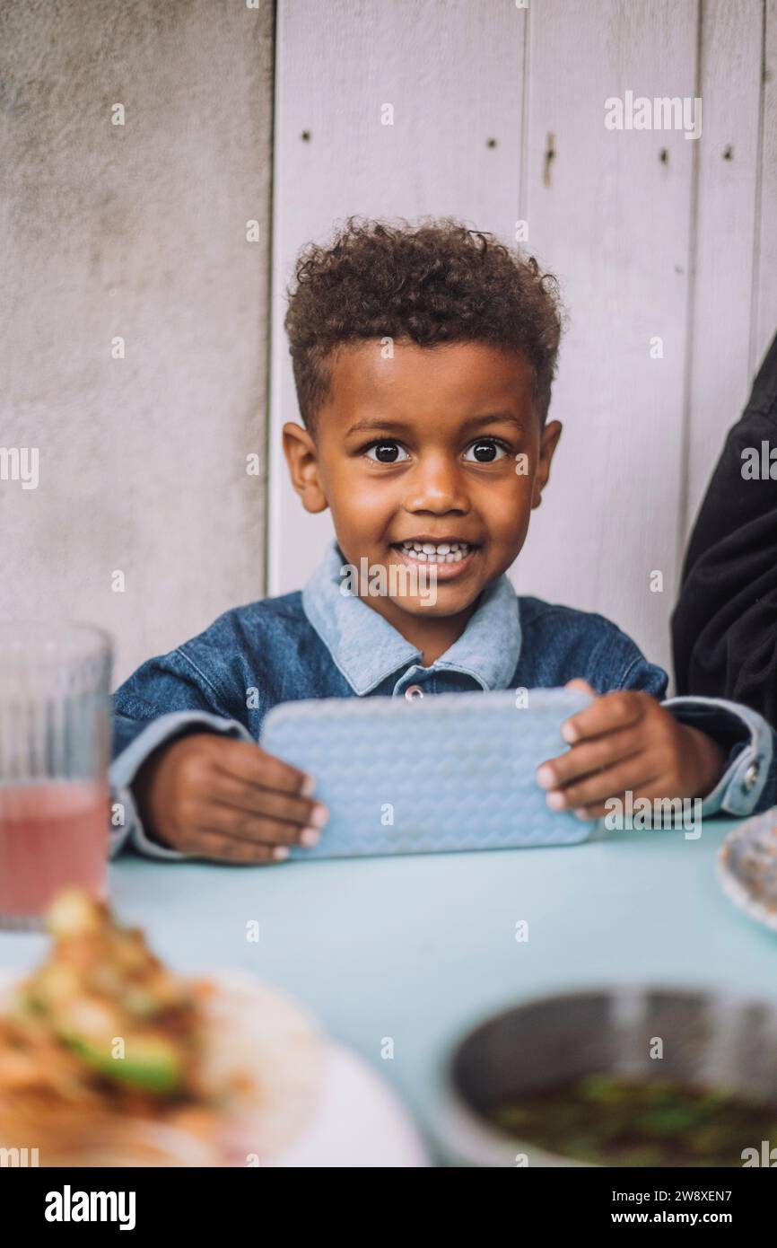 Ritratto di un ragazzo sorridente che tiene in mano lo smartphone mentre è seduto al ristorante Foto Stock