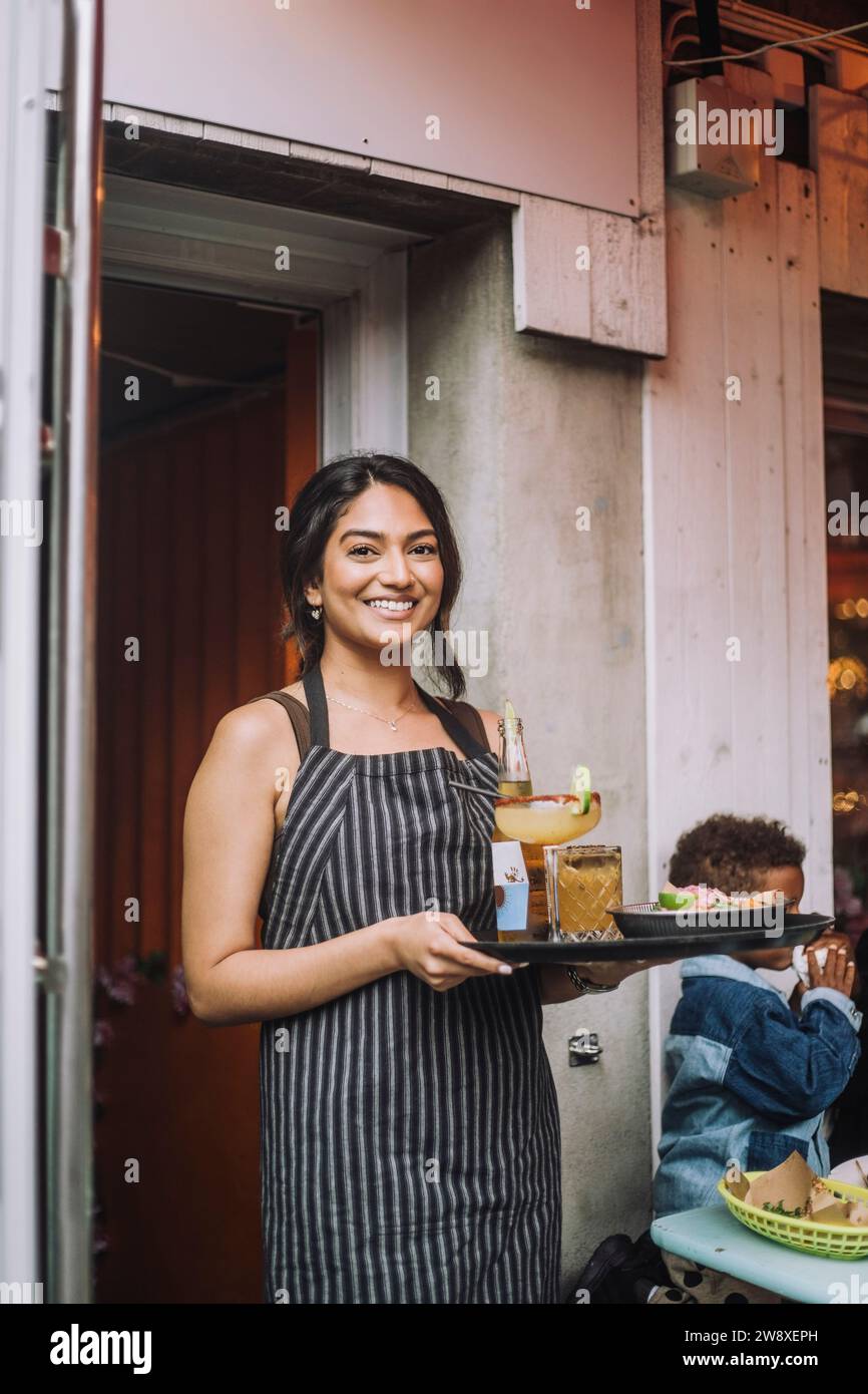 Ritratto di una cameriera sorridente che indossa un grembiule con vassoio di cibo e bevande al bar Foto Stock