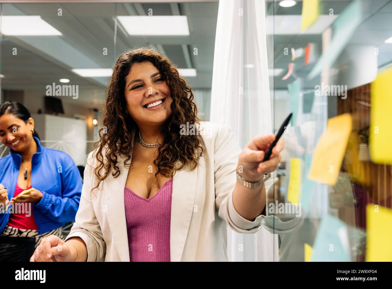 Una professionista professionista che sorride puntando verso la parete di vetro mentre si fa brainstorming con un collega in ufficio Foto Stock