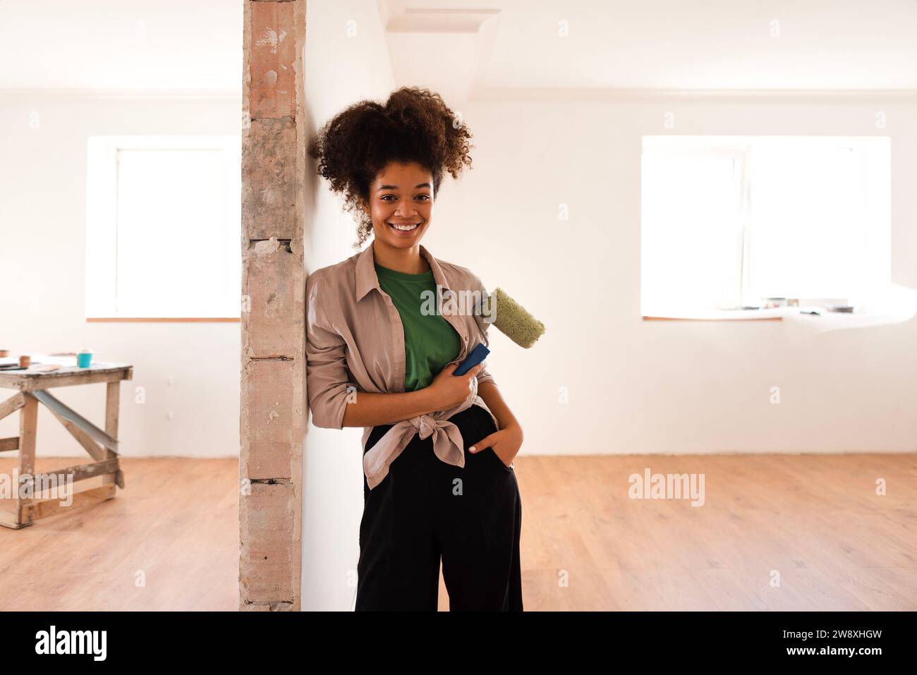 Una giovane attraente donna afroamericana in abiti casual in piedi e sorride con un rullo di vernice. Dipingere le pareti. Ristrutturazione di una casa o di un appartamento Foto Stock