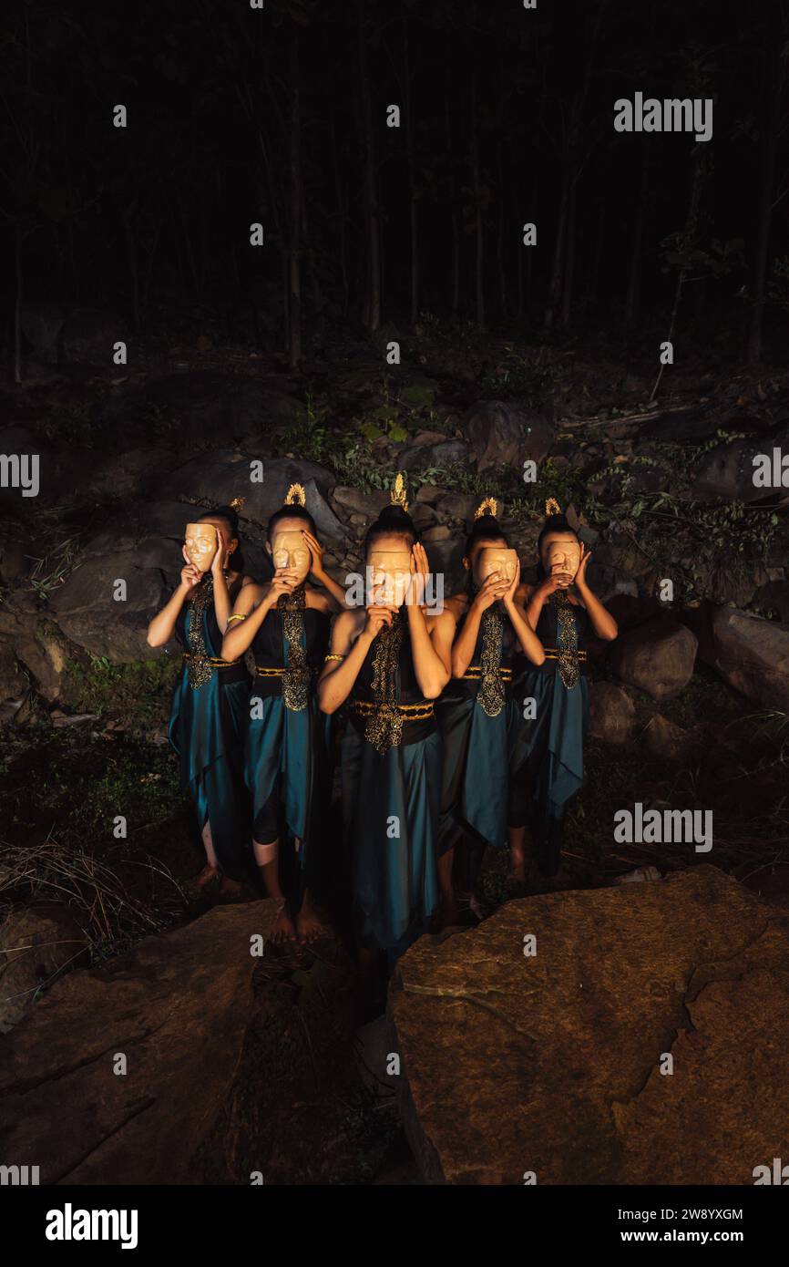 Un gruppo di donne indonesiane che tengono e indossano una maschera di legno marrone in una gonna verde tra la roccia all'interno del bosco Foto Stock