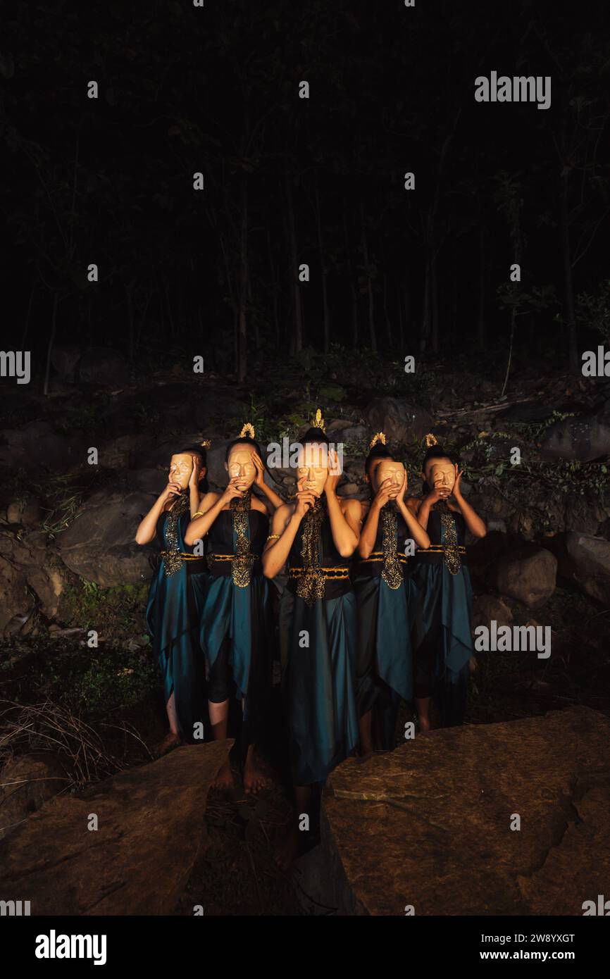 Un gruppo di donne indonesiane che tengono e indossano una maschera di legno marrone in una gonna verde tra la roccia all'interno del bosco Foto Stock