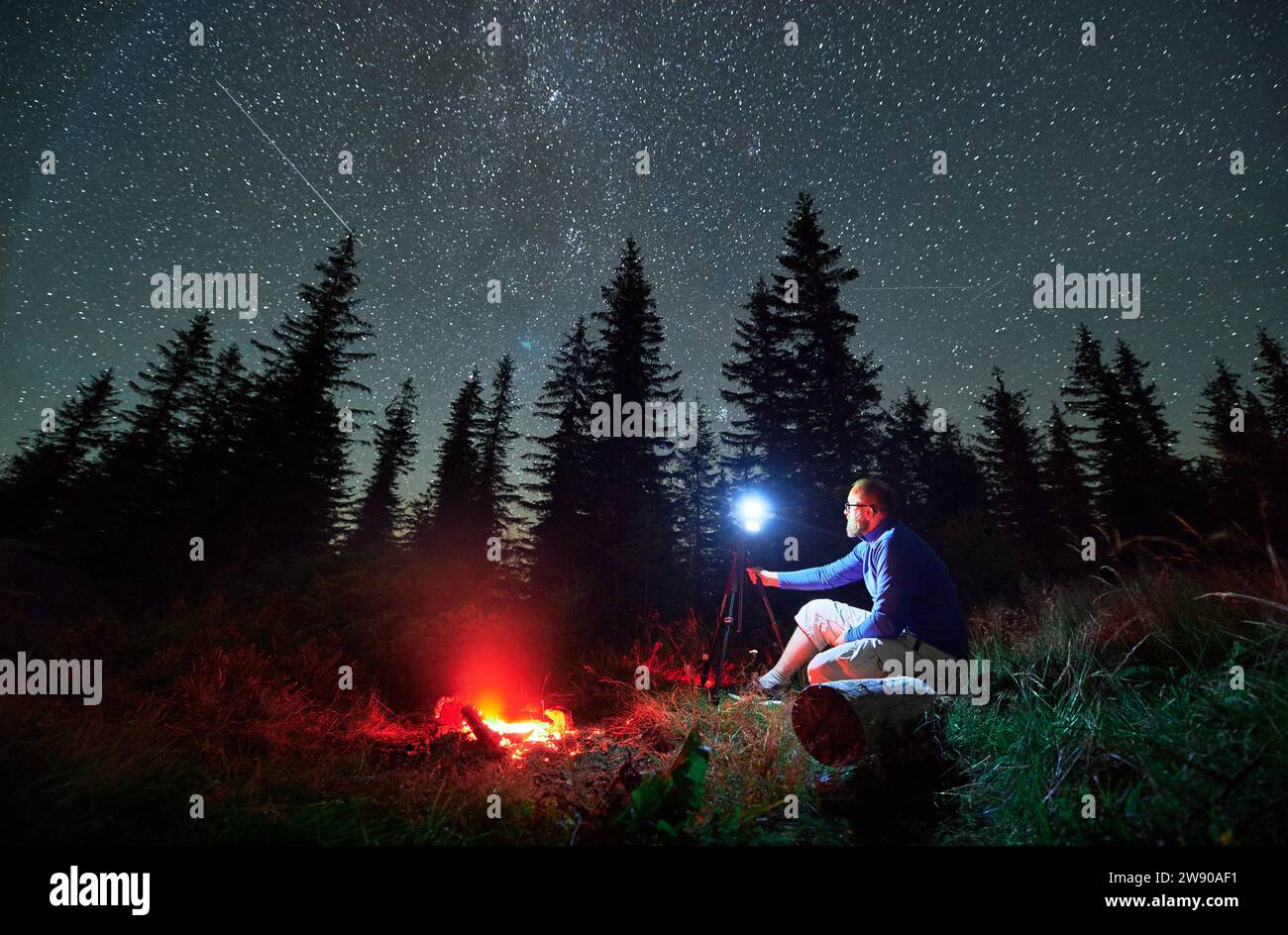 Fotografo di sesso maschile che fa servizio fotografico in montagna. Uomo che scatta foto di una notte stellata. Giovane seduto sul tronco accanto al falò di notte. Foto Stock