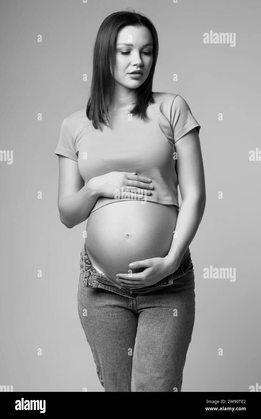 Ritratto in bianco e nero di una giovane donna incinta in t-shirt e jeans su sfondo grigio. Pancia femminile esposta. sesto mese di gravidanza. Foto Stock