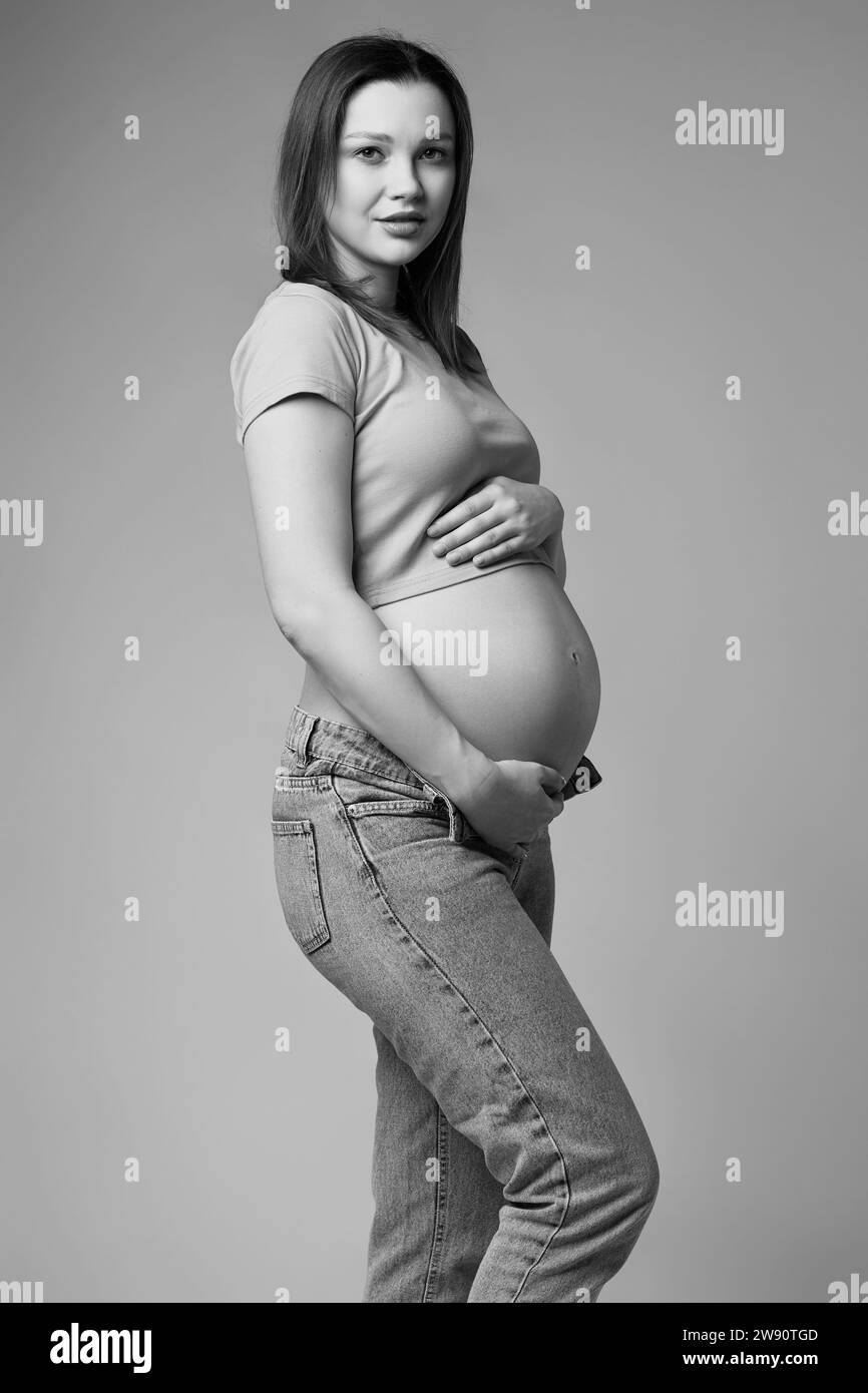 Ritratto in bianco e nero di una giovane donna incinta in t-shirt e jeans su sfondo grigio. Pancia femminile esposta. sesto mese di gravidanza. Foto Stock