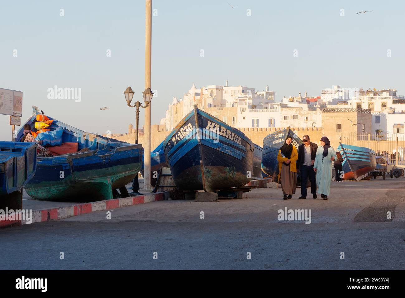La famiglia passeggia davanti ai pescherecci blu in una serata invernale con la medina alle spalle, a Essaouira, Marocco. 22 dicembre 2023 Foto Stock